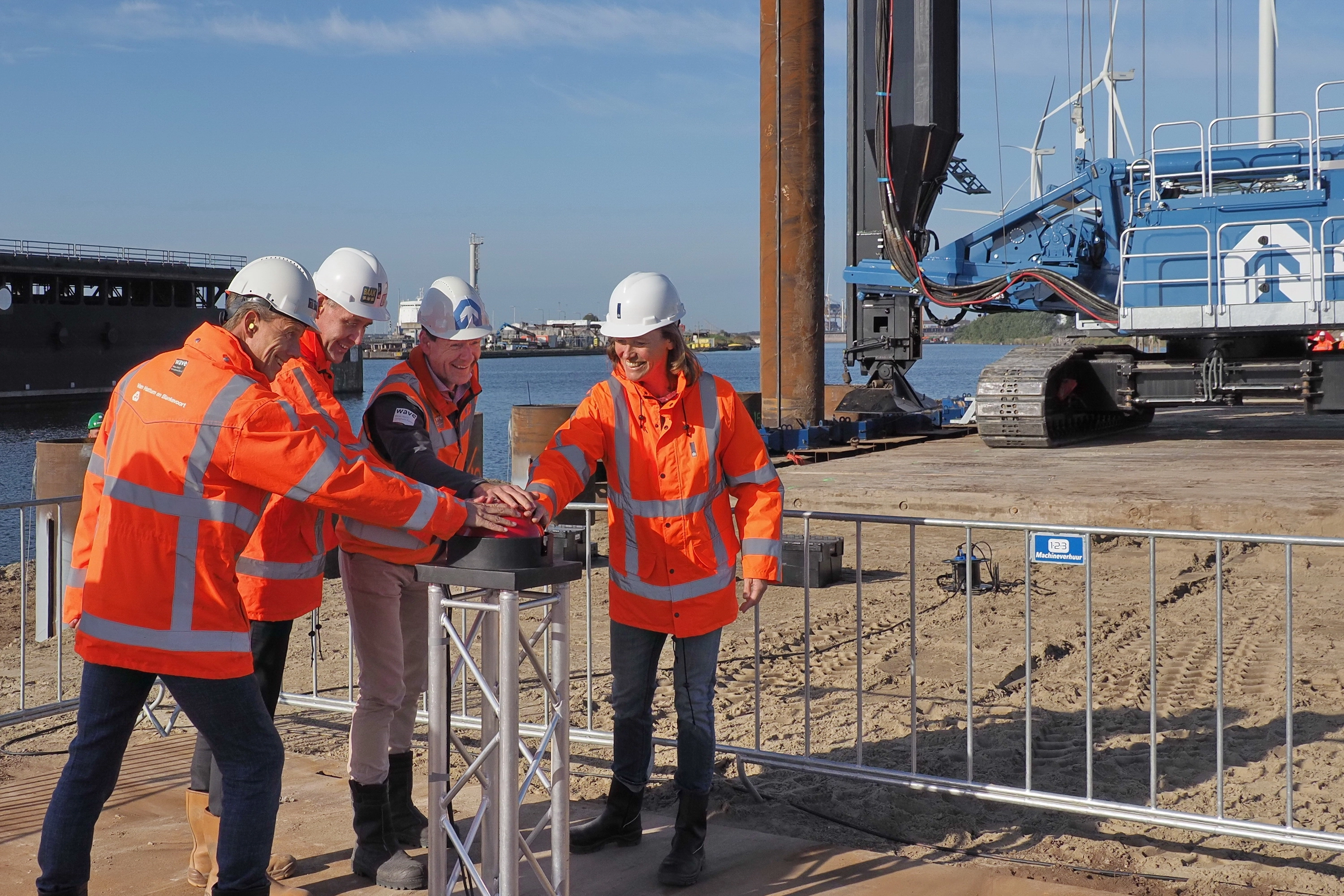 Vier mensen in oranje veiligheidskleding en helmen drukken gezamenlijk op een knop tijdens een officiële bouwstart bij water en zware machines.