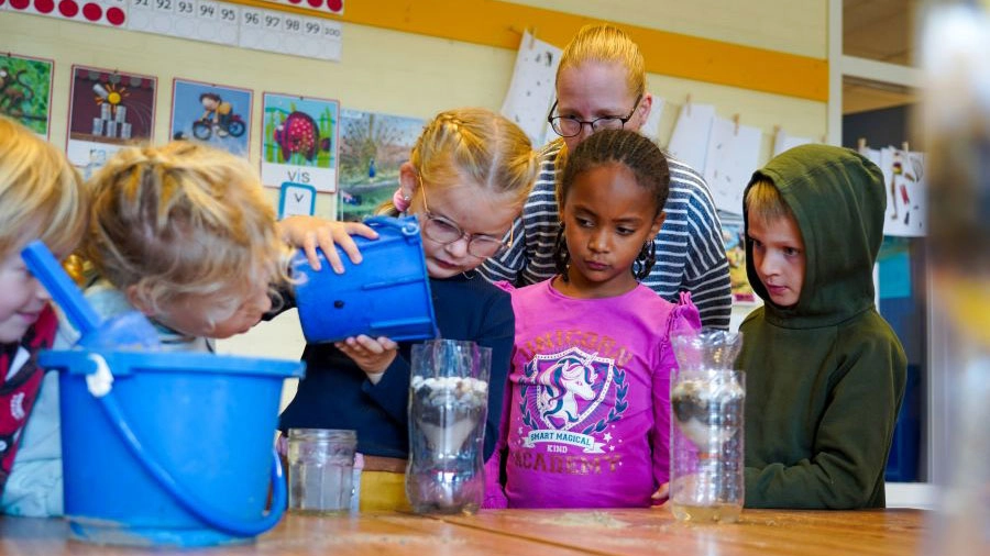 Jonge kinderen experimenteren met waterfiltratie in de klas, gebruikmakend van plastic flessen en blauwe emmers.