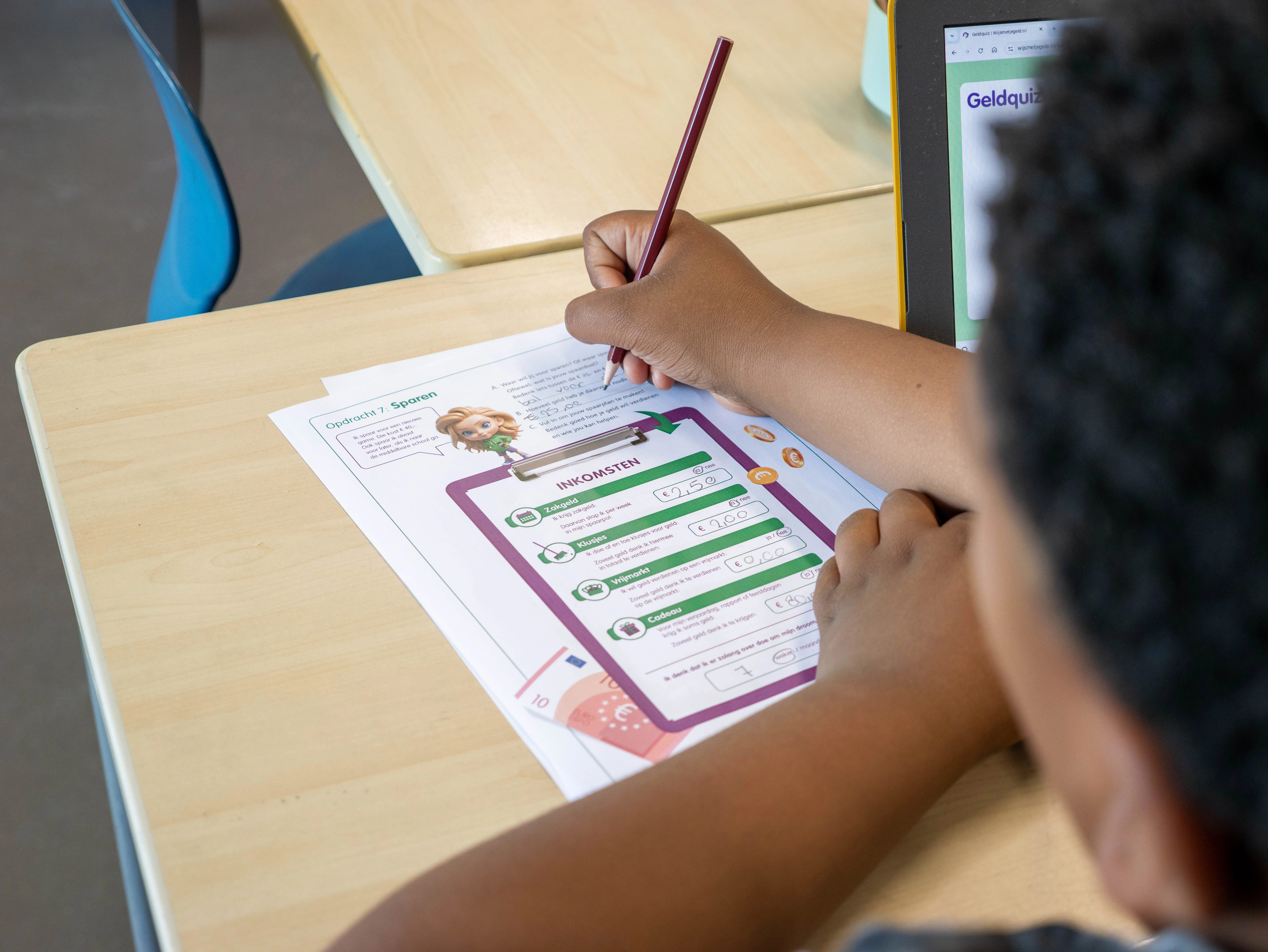 Leerling werkt aan een geldopdracht op papier met een tablet erbij, als onderdeel van lesmateriaal over financiële vaardigheden.