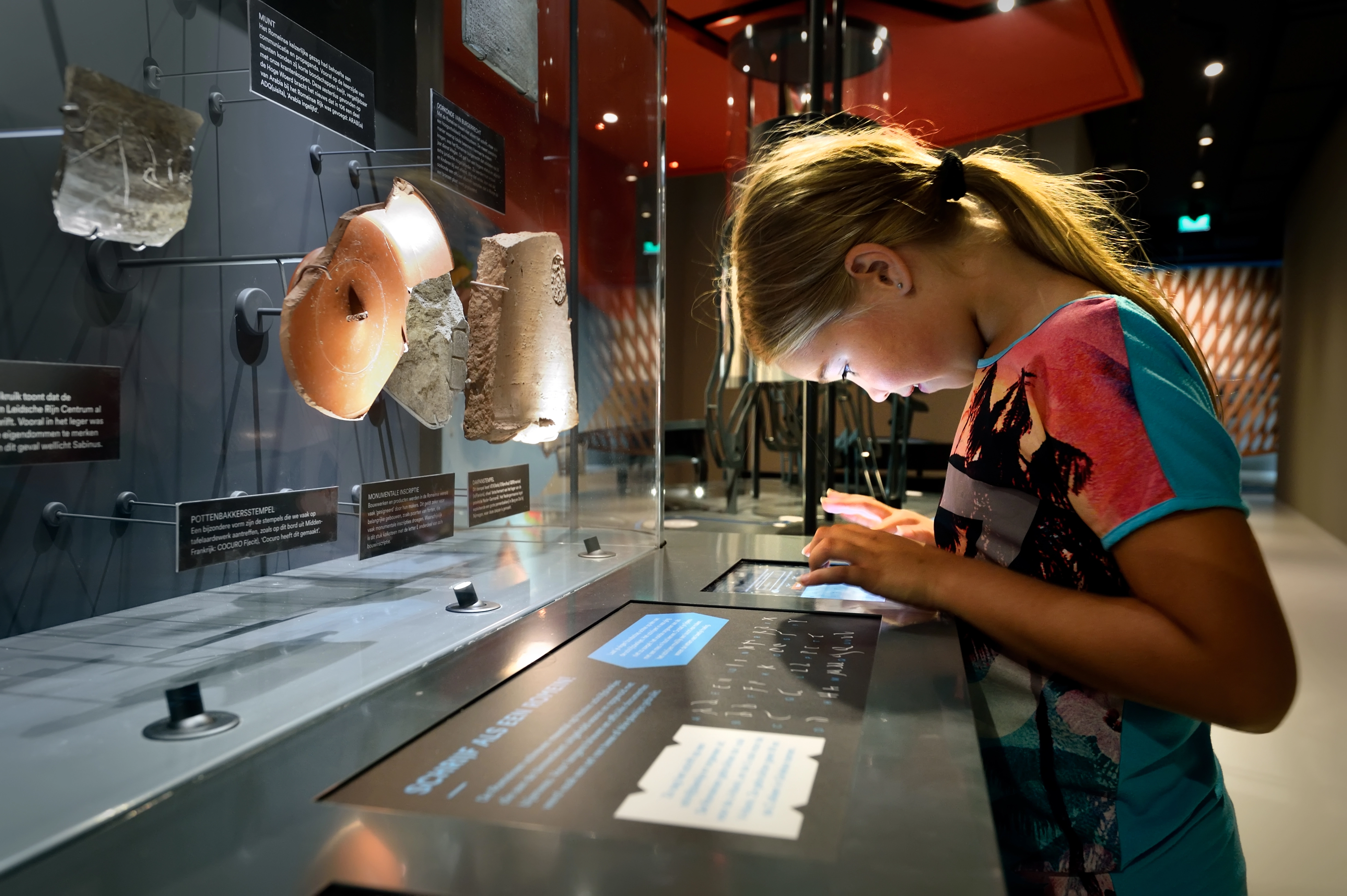 Kind onderzoekt archeologische voorwerpen en leest uitleg op tablet in museum Castellum Hoge Woerd.