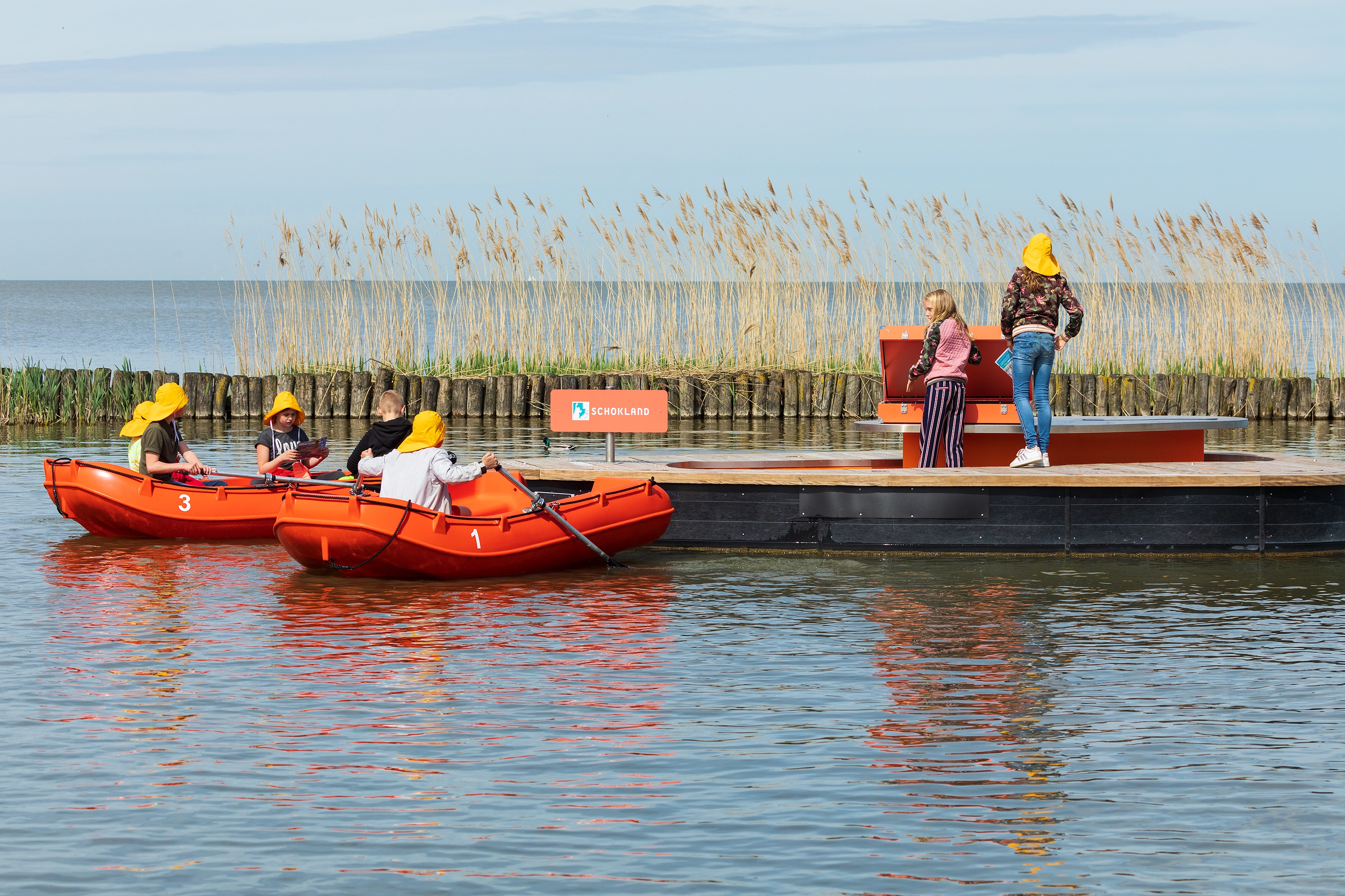 Kinderen in oranje roeiboten naderen een vlot met het bord “Schokland”, onderdeel van een speurtocht op het water.