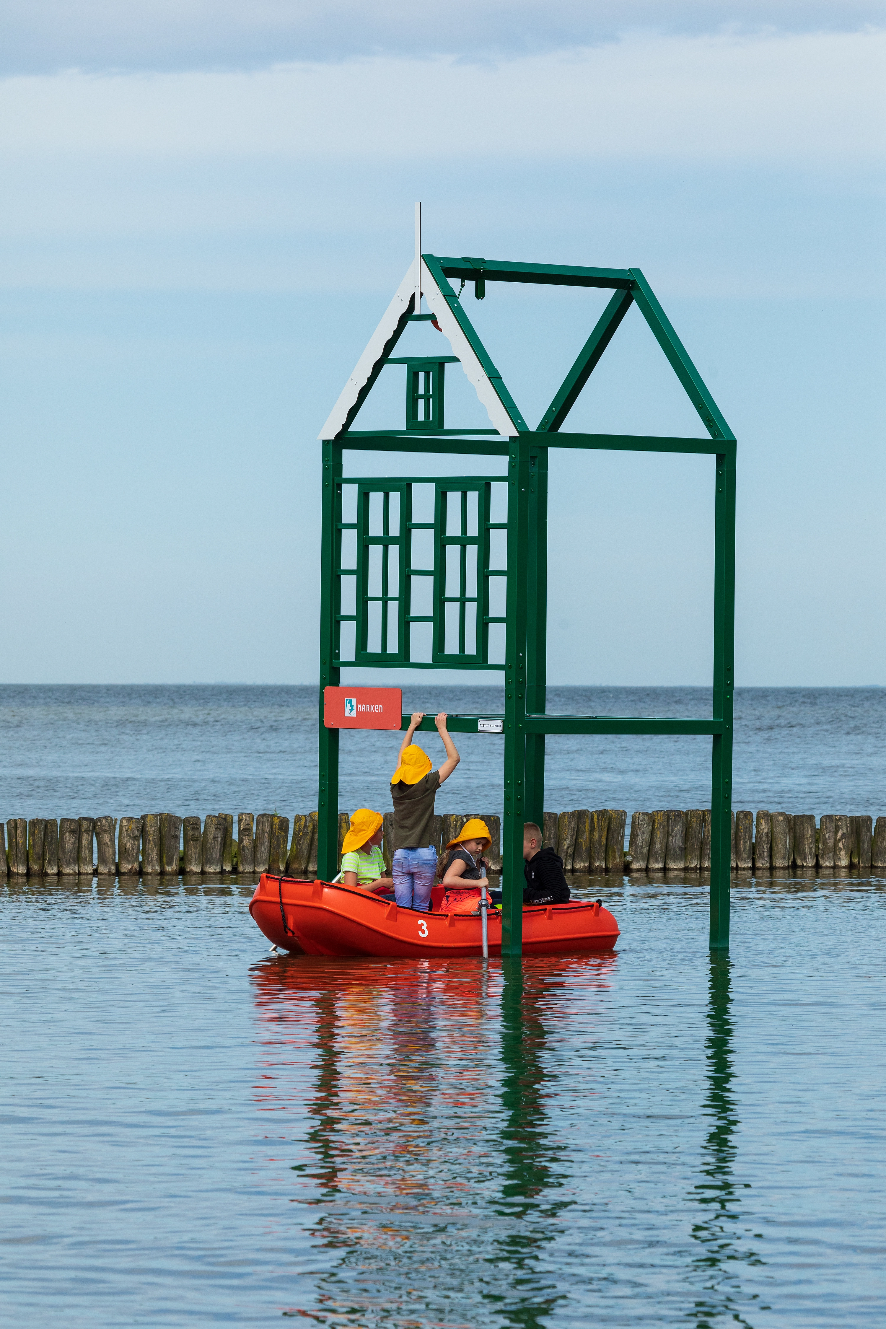 Vier kinderen in een rode roeiboot bereiken een groen metalen huisje boven het water met het bord “Marken”.