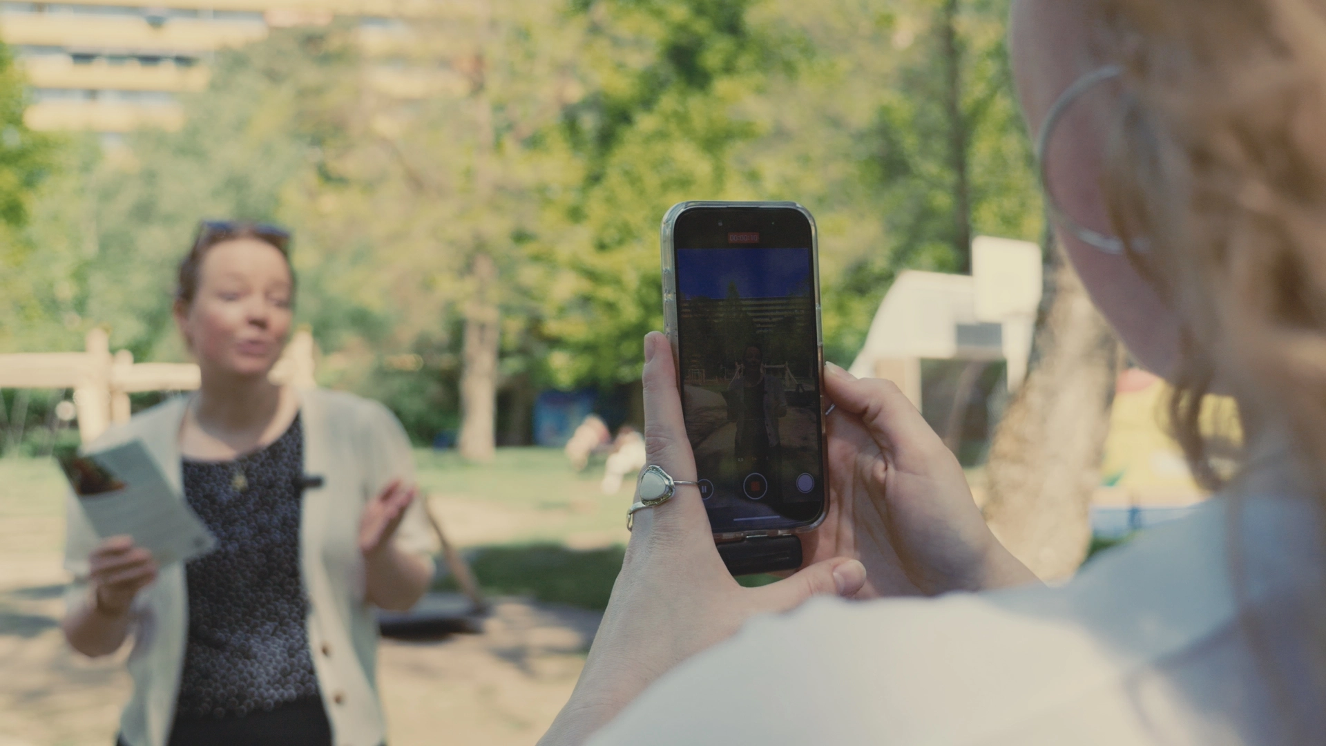 Vrouw vertelt met flyer in haar hand. Andere vrouw maakt een video van haar met een smartphone.